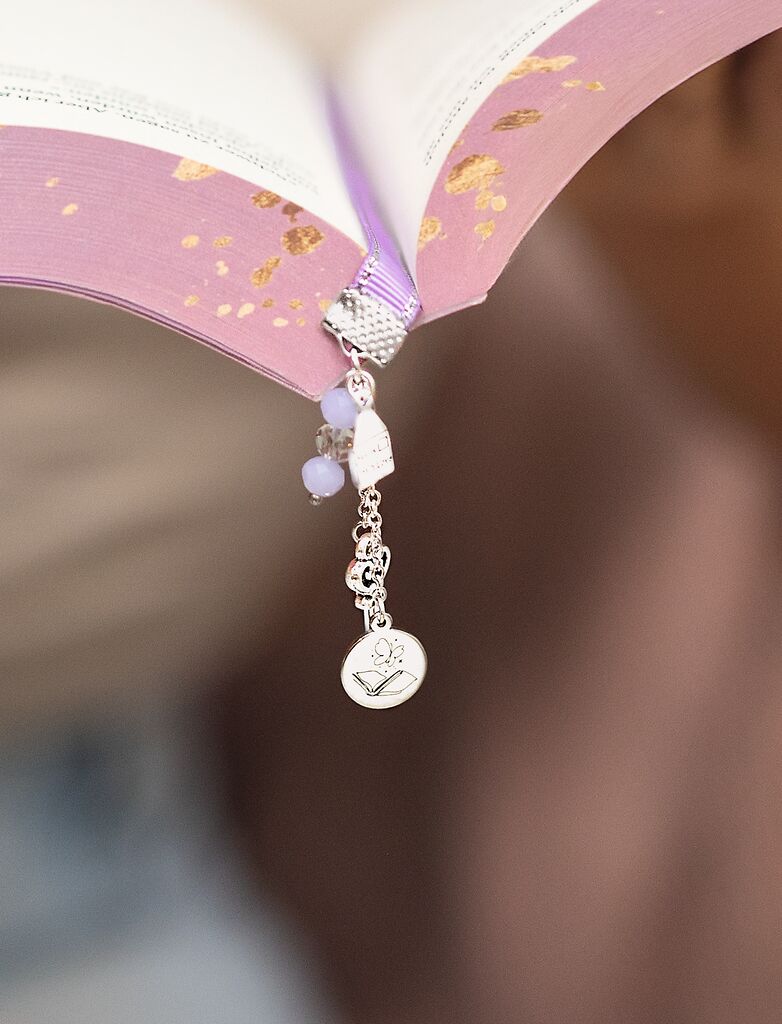 Nahaufnahme eines aufgeschlagenen Buches mit lila-goldenem Einband und einem dekorativen metallischen Lesezeichen mit einem Anhänger, Perlen und einem runden Anhänger mit Blumenmuster, der am Buchrücken hängt.